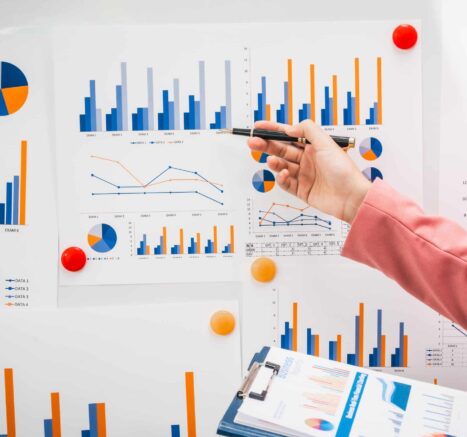 Visualize Data A hand in a pink sleeve pointing with a pen at a board filled with various charts and graphs, illustrating a leader as coach analyzing data and guiding a team.