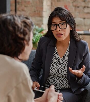 A senior executive woman A senior executive in a suit passing a leadership badge to a younger professional, symbolizing C level succession in a modern office environment.