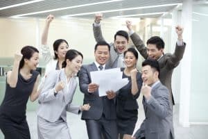 A leader in a suit stands among a team celebrating with raised fists, holding documents, depicting a leader as coach in an enthusiastic office atmosphere.