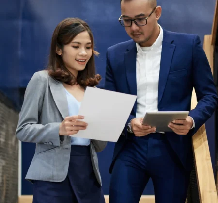 Our Team Dua profesional muda berdiskusi sambil memegang dokumen dan tablet, merepresentasikan kerja tim bersama ICF-Certified Coaches.